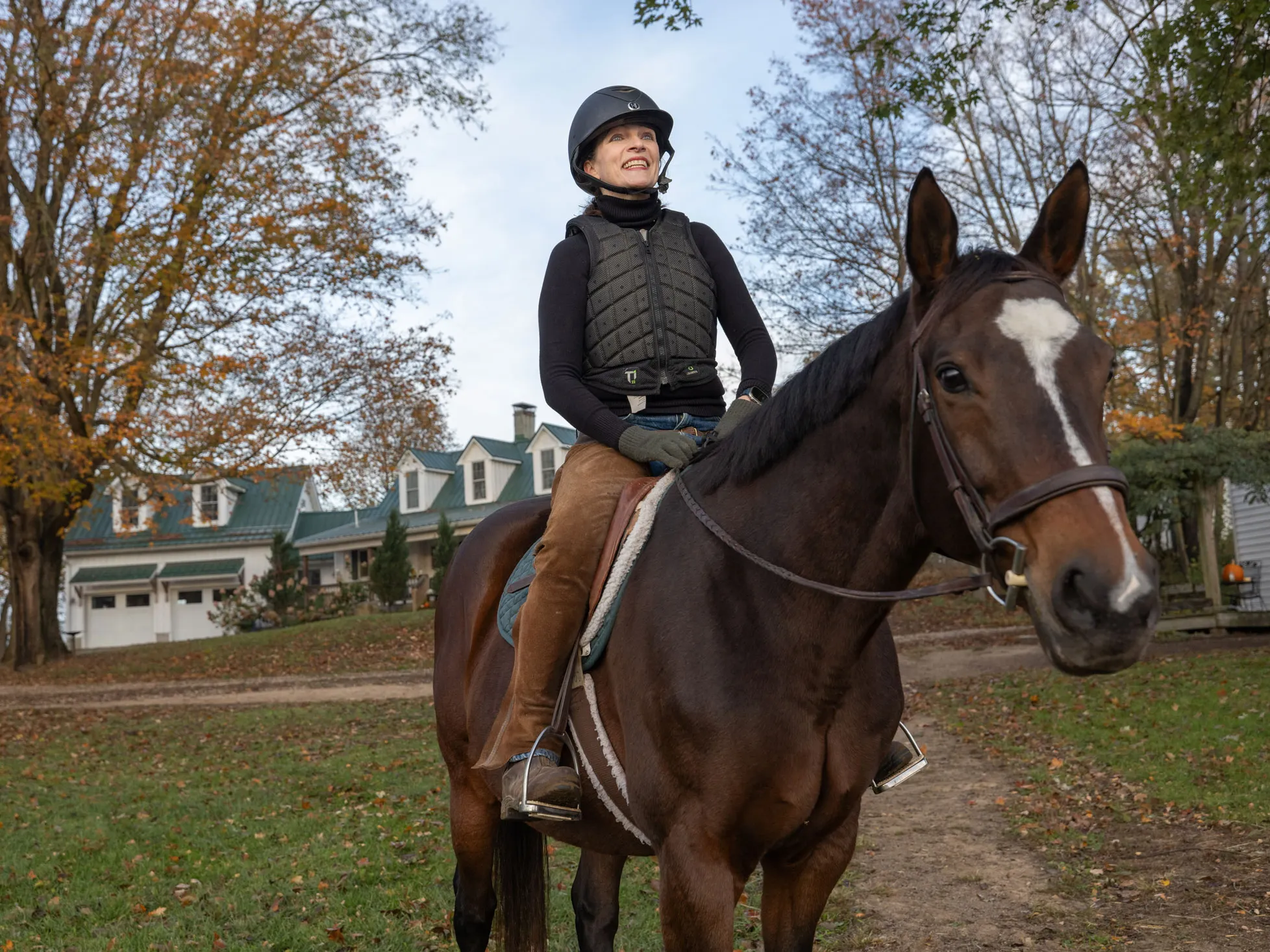 Kathy Lloyd smiles as she sits on a horse outside her riding stables on a crisp autumn day. She wears a helmet and protective riding vest sits and the horse looks directly at the camera as she looks off into the distance.