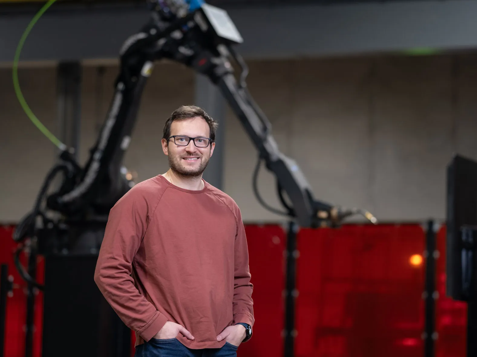 A white man smiles and stands inside a factory with his hands in pockets. Behind him is a giant robotic arm. He wears a sweatshirt, glasses and has neat hair and beard.