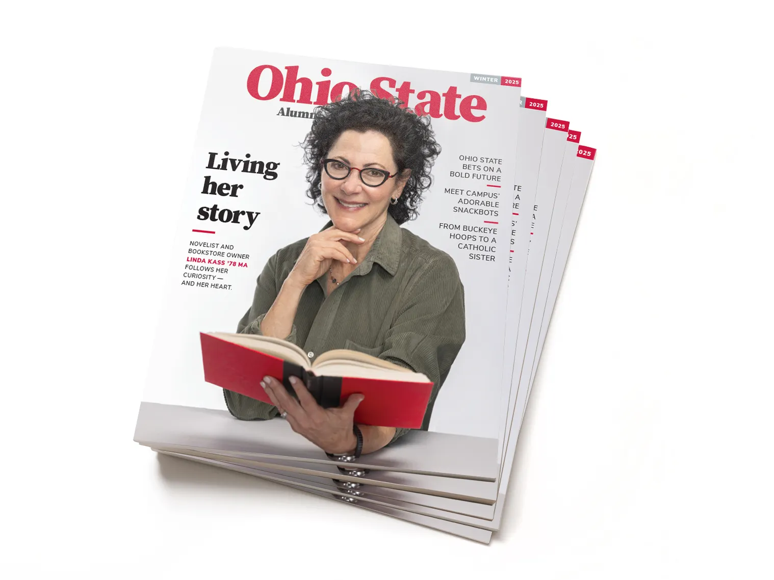 This is a stack of the Winter 2025 edition of Ohio State Alumni Magazine. The cover shows alumnus Linda Kass holding a book. Her other hand is tucked just beneath her chin. She’s a white woman with curly dark hair and glasses, and she looks happy, smart and well put-together. Her story is titled “Living her story” and other text on the cover says “Ohio State bets on a bold future”; “Meet campus’ adorable snackbots”; and “ From Buckeye hoops to a catholic sister”