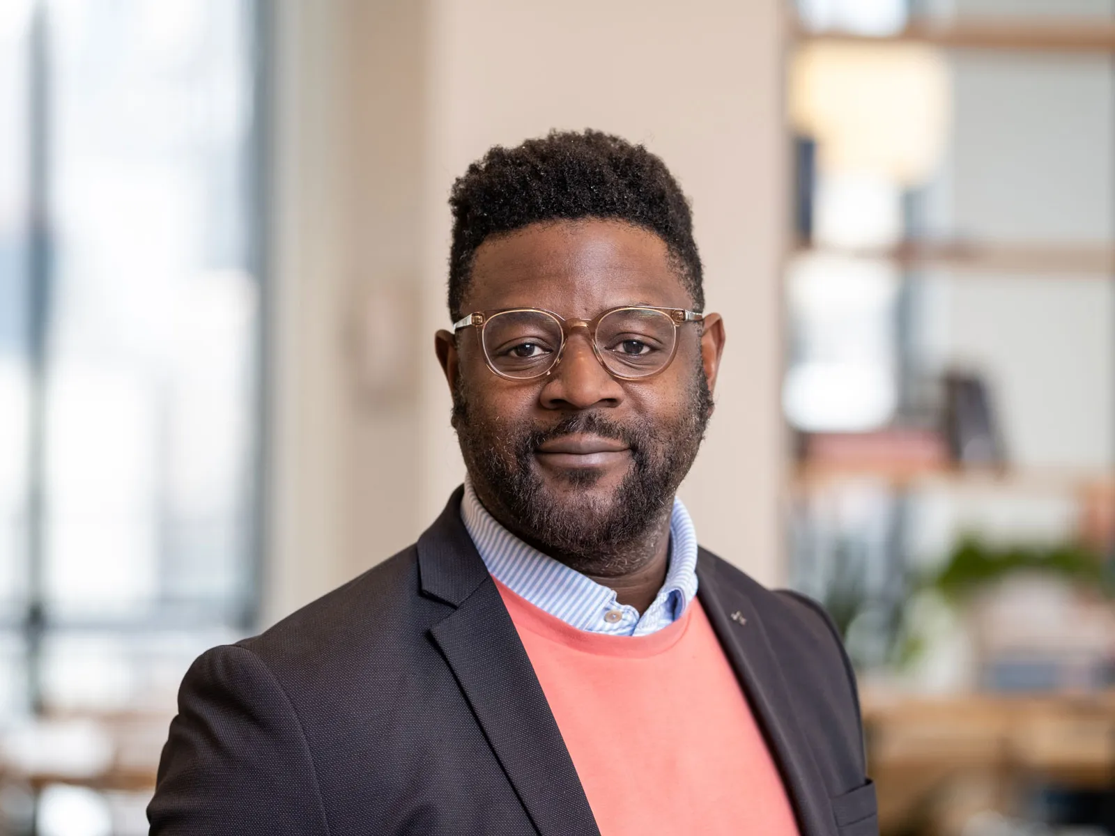 A Black man slightly smiles as he poses for this portrait in a modern office setting with shelves and large windows in the background. He wears glasses and sport coat over over a collared shirt and sweater.