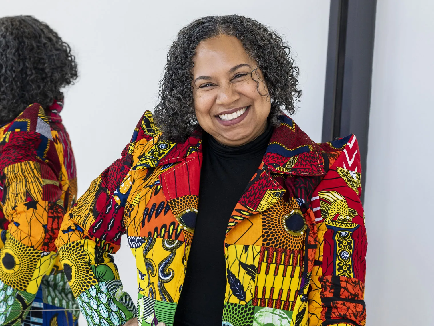 A Black woman grins as she holds a closed laptop and stands in front of a large mirror. IT reflects the back of her curly hair and patterned, stylish jacket. The hand not holding her laptop is on her hip. She comes across as bright, sunny and well-styled.