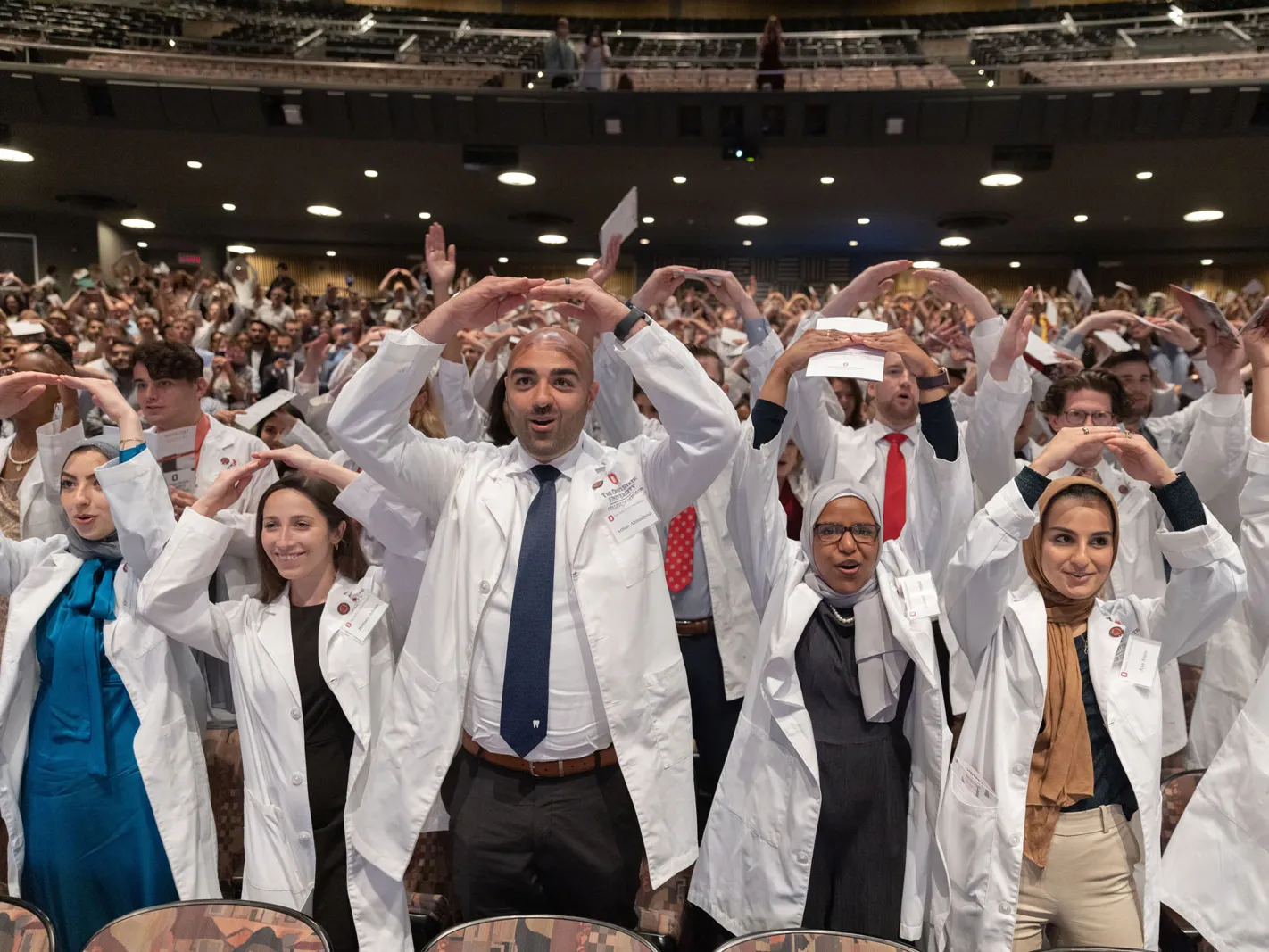 An auditorium is filled by College of Dentistry students wearing white coats, standing up from their seats, and holding their arms rounded above their heads so their fingertips touch. They’re making an O for the Buckeyes’ O-H-I-O cheer. 