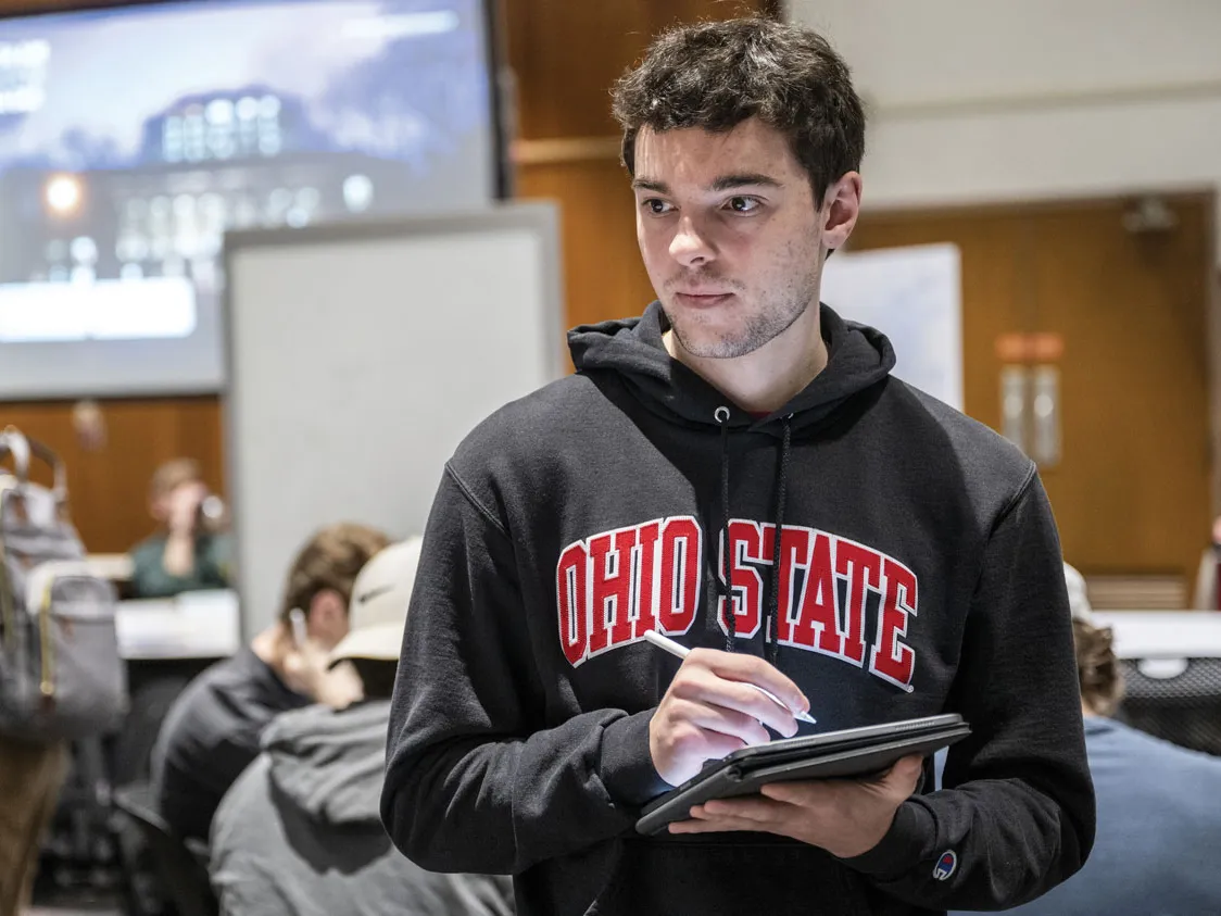 A student takes notes as classmates talk about his work. He wears an Ohio State sweatshirt and an intensely focused expression