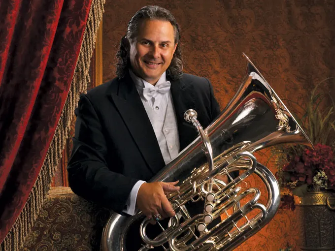 In his younger days, a long-haired Jim Akins smiles as he poses for an official portrait wearing a tux and holding his shiny tuba. A fancy background includes a velvet drape and brass pot of flowers.