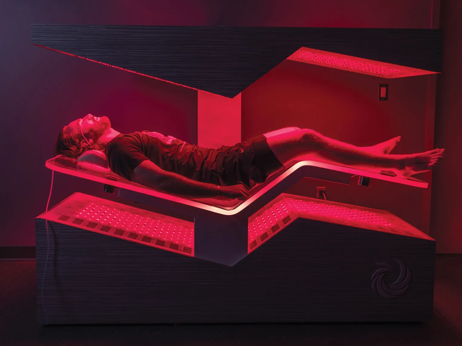A man lies in a futuristic bedlike machine. The machine surrounds his body above and below in a structured, enclosed shape. His legs are slightly elevated and his arms rest beside his torso. Red light shines over him.