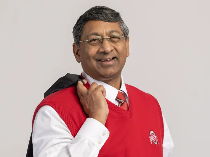 Ravi Bellamkonda smiles as he holds his suit coat flipped over his shoulder. He wears a scarlet sweater vest, plaid tie and glasses. 