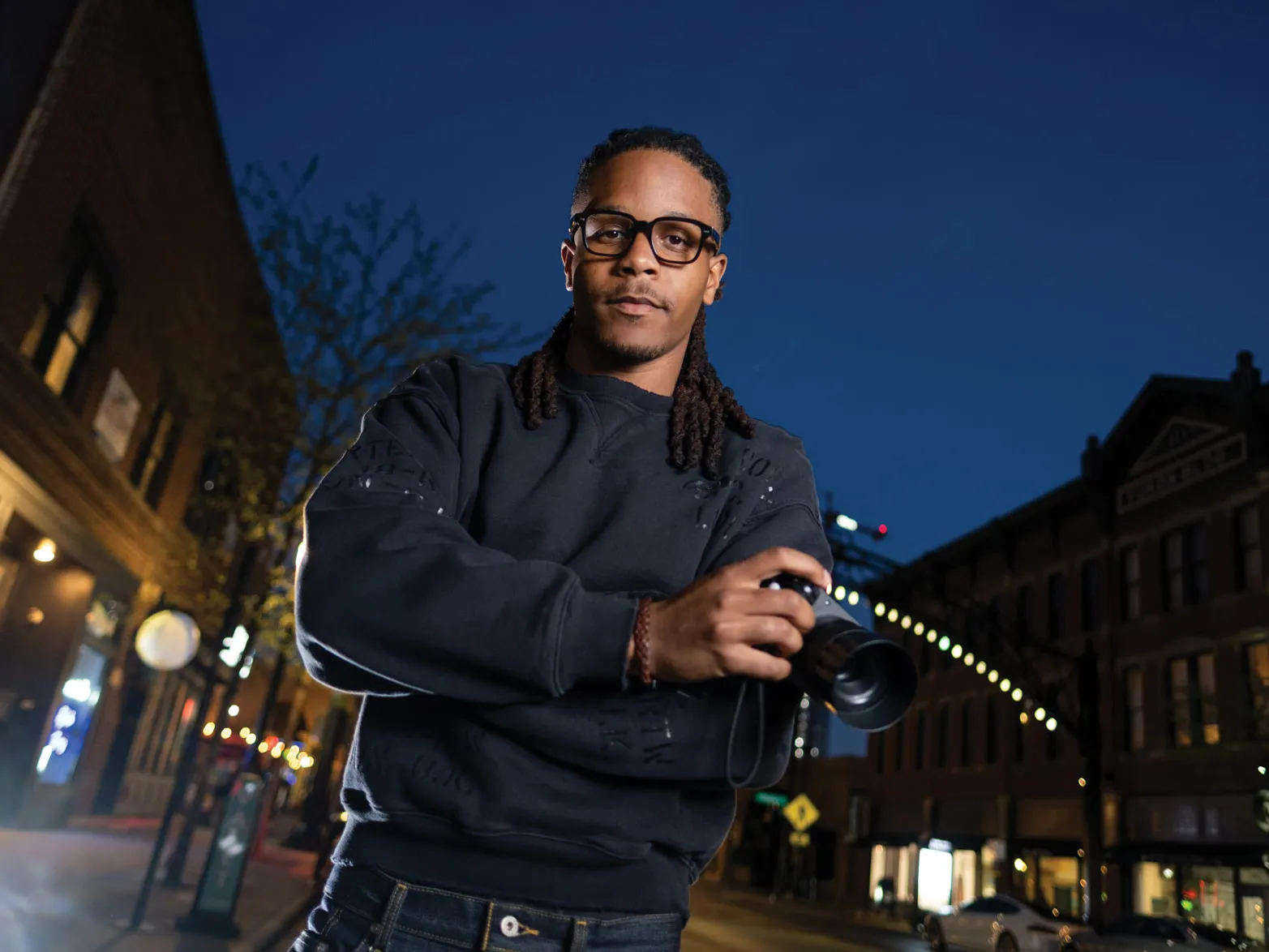 Justen Turner is a young black man wearing thick-framed glasses and a sweatshirt. He looks focused as he poses for this portrait holding a small video camera. His hair is dreads pulled back in a low ponytail. He looks like he's competent, professional and thoughtful—or like someone you'd trust to make a movie about yourself or a loved one. He's photographed in the Short North after the sun has set.