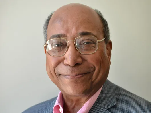 Noel Mayo smiles for a portrait, wearing glasses and a checked sport coat, but no tie.
