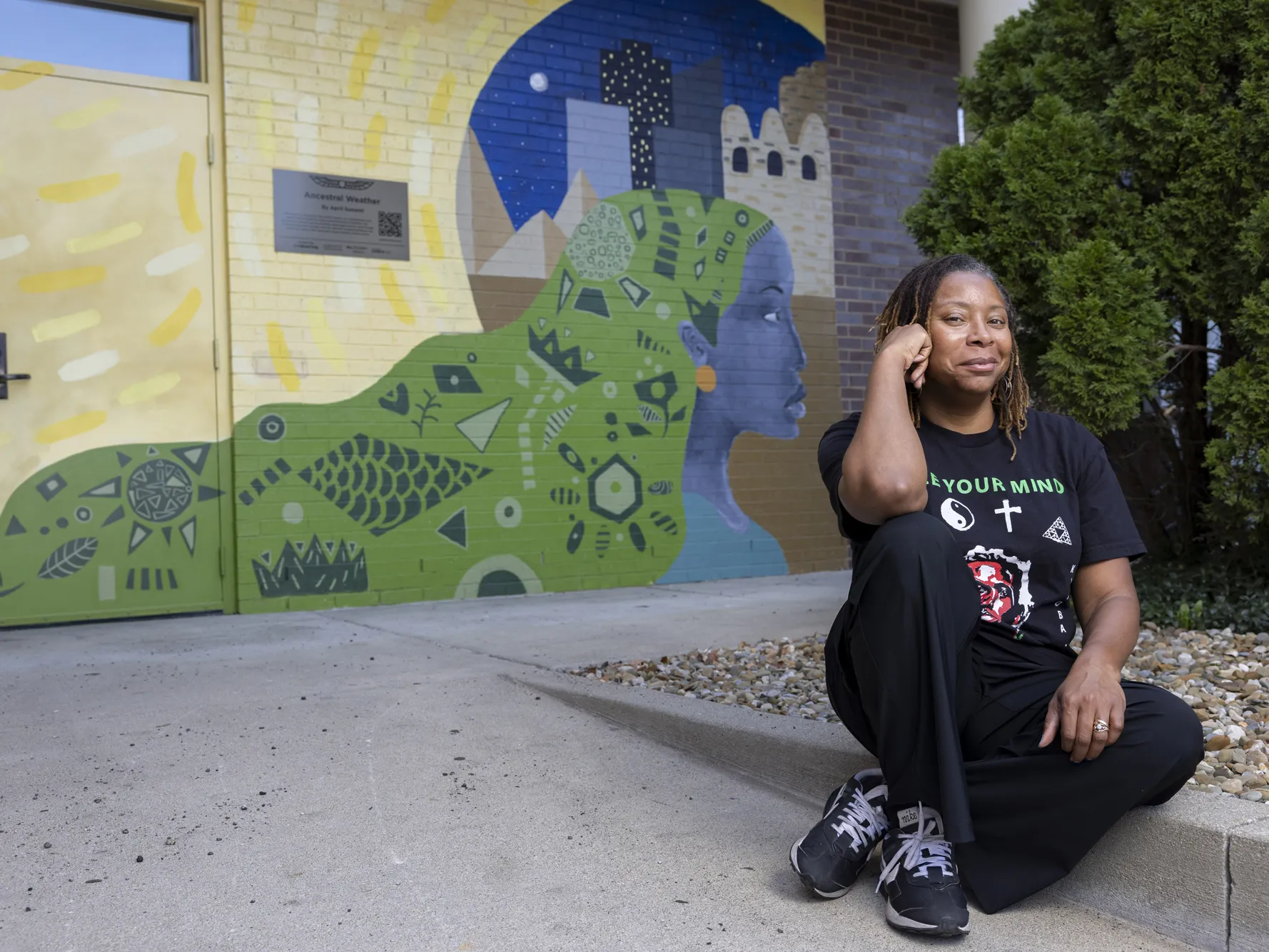 April Sunami smiles knowingly as she sits on a curb in front of a colorful mural she painted