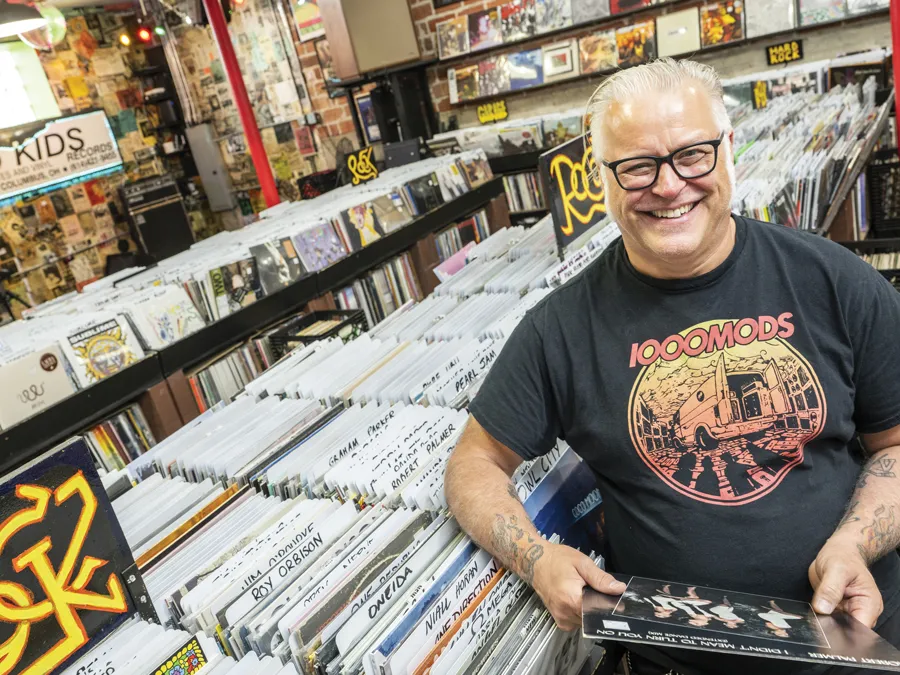 Used Kids owner Greg Hall laughs as he is photographed in the record store, wearing thick-framed glasses and an artful T-shirt for the hard rock band 1000 Mods.